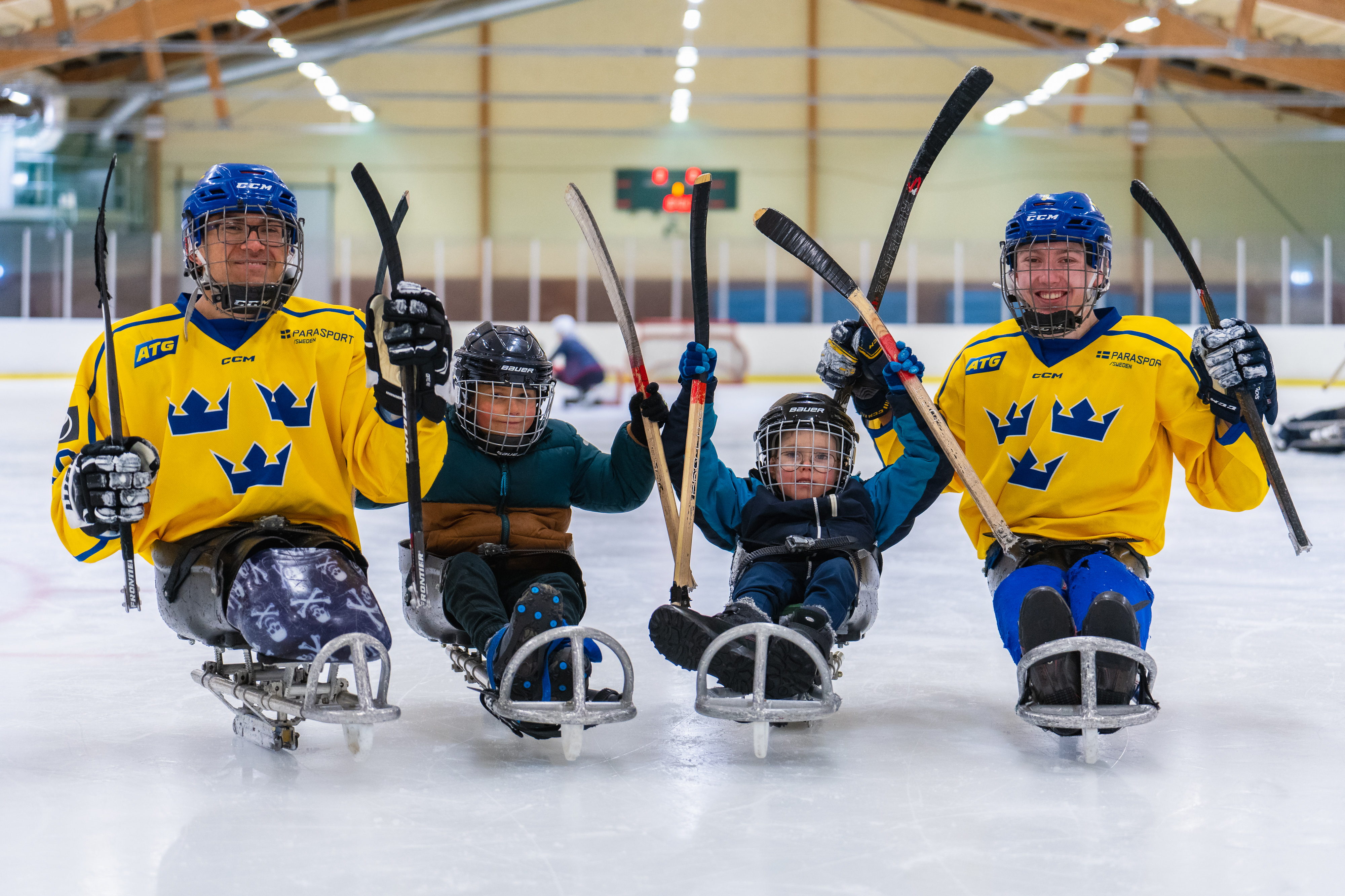 Inomhus, ishall. Fyra paraishockeyspelare. Två barn som provar på i mitten, på vardera sida en äldre landslagsspelare. De sitter på isen i kälkar med utrustning och hockeyklubbor. 